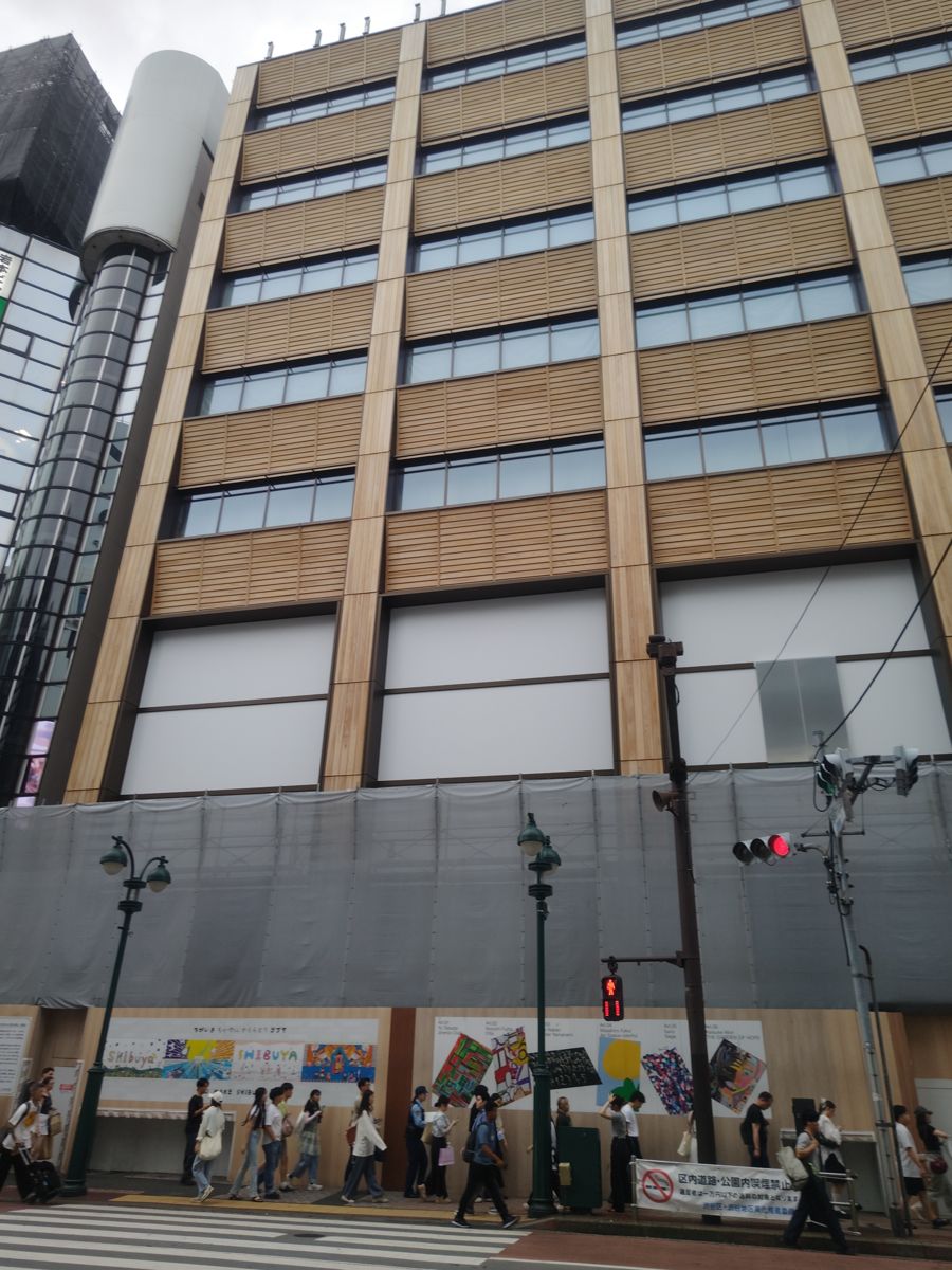 建設中の9階建てのビル。各階の窓のあいだに木色の縦柱と横板が並び、1階まわりはシートと仮囲いで覆われている