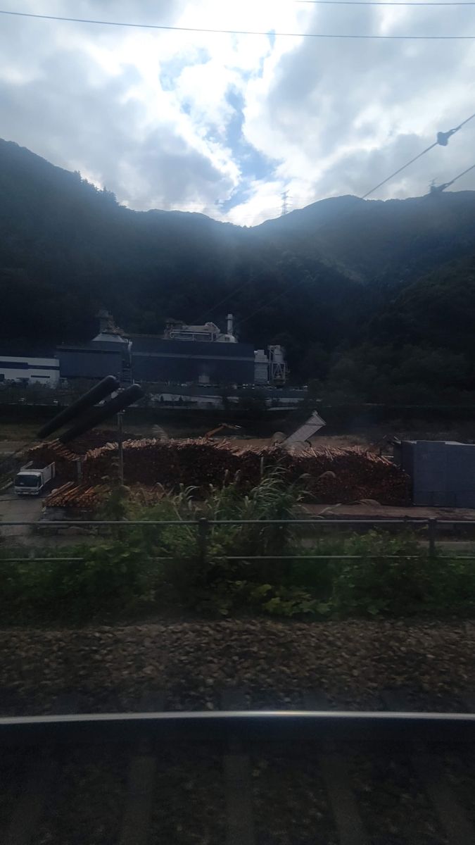 電車の窓から見た、山の手前に積み上げられた丸太の山と発電所の建物