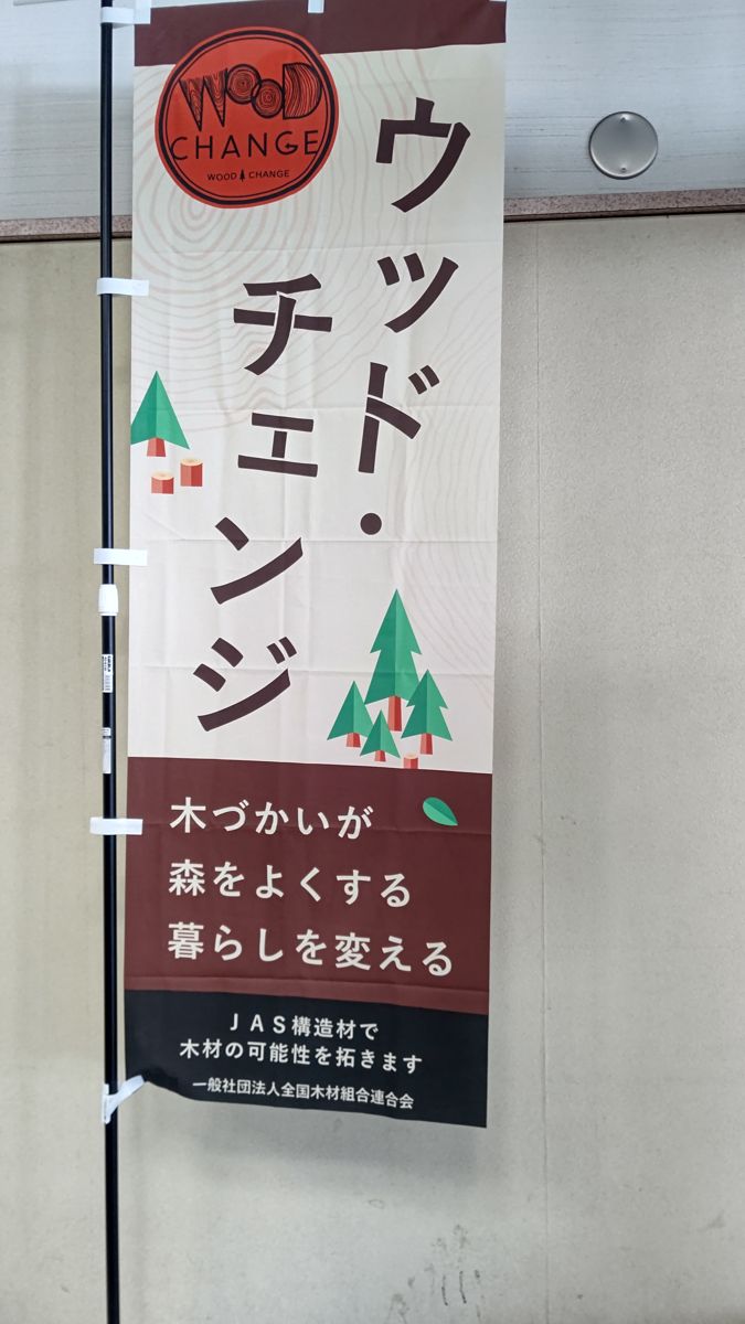 庁舎の壁の前に立つのぼり旗。「ウッド・チェンジ 木づかいが森をよくする 暮らしを変える」「JAS構造材で木材の可能性を拓きます 一般社団法人全国木材組合連合会」と書かれている
